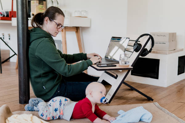 Woman working on a laptop computer, propped up on a low chair, as she also takes care of her child.
