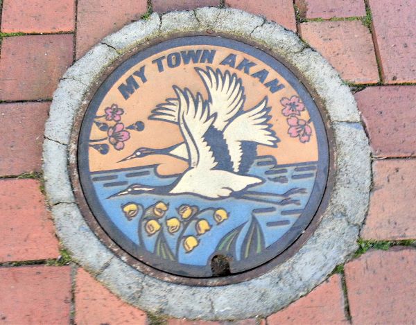 Manhole cover featuring a pair of airborne red-crowned cranes over wetland, surrounded by pink sakuras and yellow snowflakes.