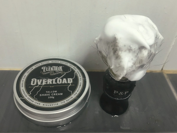 Close up of a tub of The Elektrik Chair Overload shaving cream, and next to it, a shaving brush that's loaded with the lather.