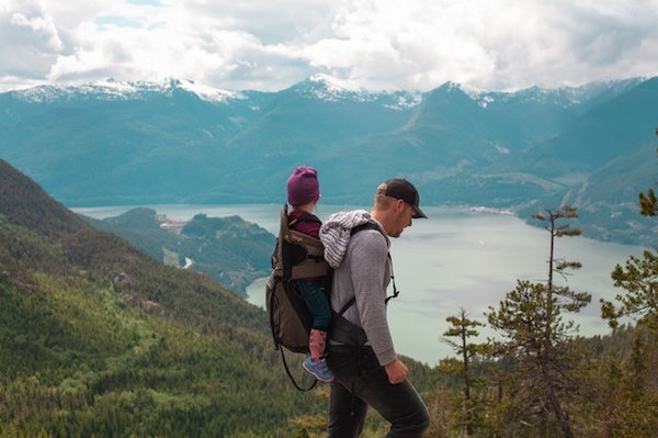 A man carrying his young daughter in a back harness on a hike around a mountain and river to venture in their own adventure race.