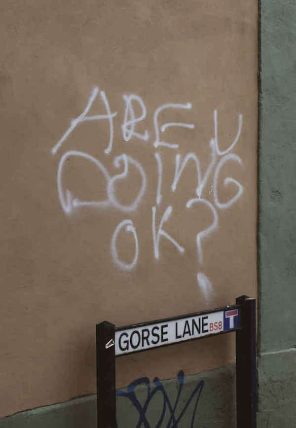 A graffiti on a wall saying "Are u doing OK? above a road sign that reads "Gorse Lane". 
