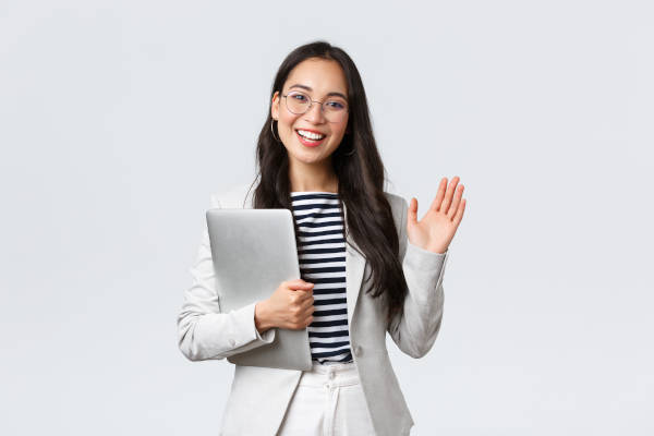 A smiling woman dressed in grey, white, and black corporate clothes waves "hi" to the camera. Her overall appearance and likeable demeanour boosts her image and etiquette skills.