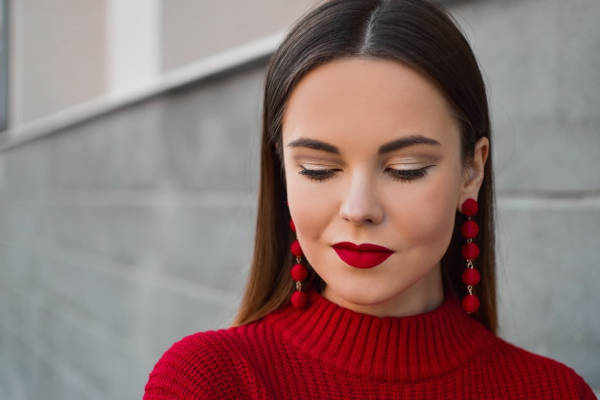 A woman dressed in red with matching red earrings and red lipstick. She glances downwards, showing a clear view of her face makeup.