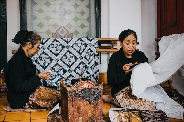 Two women sitting down and making batik using canting. 