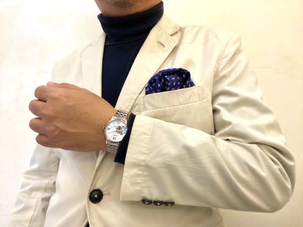 An off-white, cotton sport jacket, navy roll-neck jumper, and a navy, small-patterned pocket square. Orient Clover on the wrist, showing how to pair a typically ladies' watch with classic menswear.
