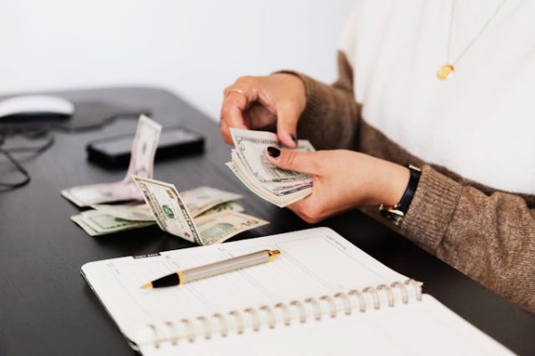 Close-up of a pair of hands counting money on a table. Also visible are a notebook, a pen, a smartphone, and a computer mouse. This symbolises the cost of home ownership.