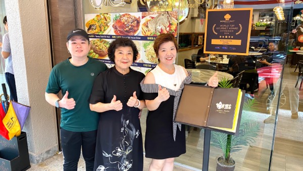 Group photo of Vincent Tan (head chef), Michele Tan (face and namesake) and Agnes Chua (overall restaurant management), standing at the front door, leaning against the menu stand.