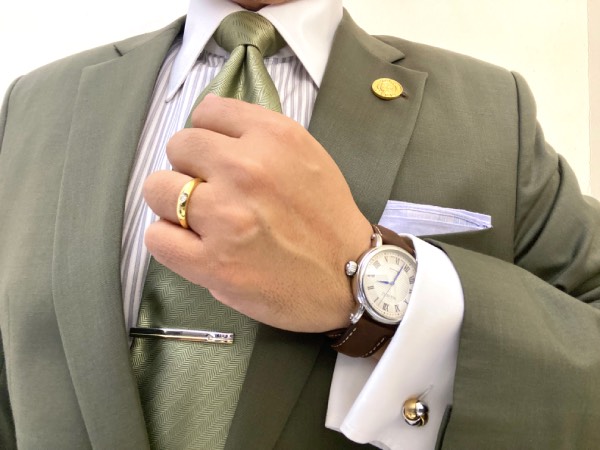 Close-up to the chest of a standard business semi-formal outfit. Image shows multiple jewellery pieces: tie clip, cuff link, lapel pin, wristwatch and wedding band, worn together in harmony, with none of them competing for attention over the other.