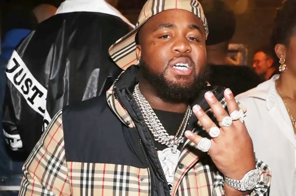 Image of rapper MO3, captured during an event, adorned with lots of jewellery. He flashes his left hand, diamond encrusted rings on each finger, watch on his wrist, and a neck full of necklaces, one of which with a huge pendant.