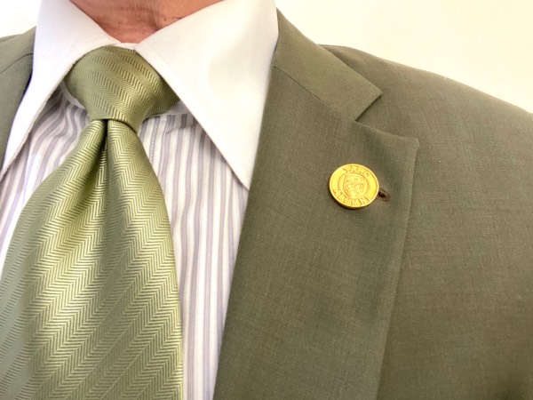 Close-up of a round, gold lapel pin, attached to the lapel buttonhole of the suit jacket. Worn with a shirt, a tie, and a pocket square.