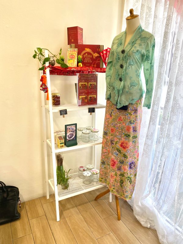 A mannequin, dressed in a light green, patterned kebaya blouse, and an overall peach sarong, with printed flowers motif in red, pink, purple and green.