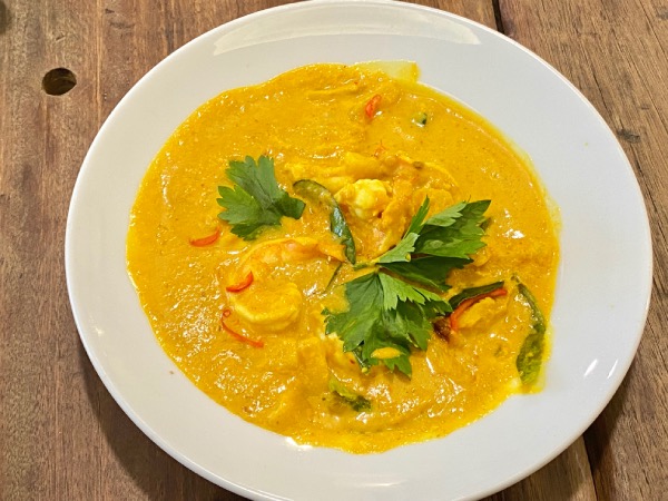 Close-up view of a plate of Sri Nyonya Bistro's Pineapple Prawn Curry. Succulent pieces of large prawns, soaking in sweet, spicy and tangy, bright yellow/red gravy. This dish is garnished with fresh parsley leaves, giving it an attractive contrast of green against an overall yellow dish.